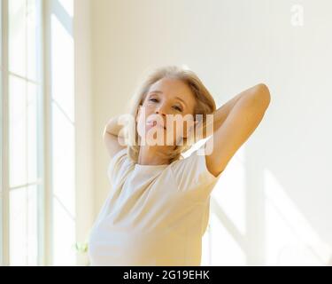 Nouveau jour. Belle heureuse femme âgée des années 60 qui se réchauffe les bras en s'étirant le matin devant la fenêtre à la maison, optimiste femme à la retraite exprimant optimisme et bonheur pendant la retraite Banque D'Images