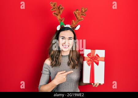 Jeune fille hispanique portant un chapeau de noël cerf tenant un cadeau souriant heureux pointant avec la main et le doigt Banque D'Images