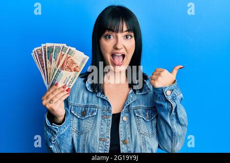 Jeune femme hispanique portant des billets en livres égyptiennes pointant du pouce vers le côté souriant heureux de la bouche ouverte Banque D'Images