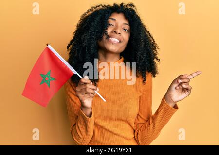 Femme afro-américaine avec cheveux afro tenant le drapeau marocain souriant heureux pointant avec la main et le doigt sur le côté Banque D'Images