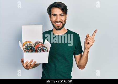 Jeune homme hispanique tenant une boîte de beignets colorés et savoureux souriant, souriant, avec la main et le doigt sur le côté Banque D'Images