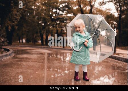 Drôle enfant fille de 3-4 ans tenant parapluie porter des bottes de pluie et manteau de pluie dans le parc marchant dans les flaques à l'extérieur. Regarder l'appareil photo. Saison d'automne. Enfant Banque D'Images