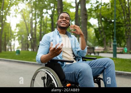 Joyeux homme noir handicapé en fauteuil roulant parlant sur smartphone au parc vert Banque D'Images