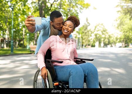 Homme noir romantique et sa petite amie en fauteuil roulant emportant le selfie ensemble, embrassant au parc de la ville Banque D'Images