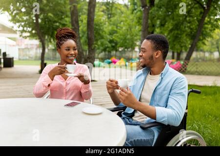 Jeune homme noir handicapé en fauteuil roulant le jour avec une jolie femme dans un café extérieur, boire un café, profiter de la conversation Banque D'Images