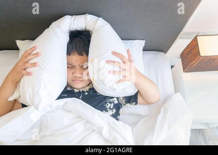 Garçon obèse allongé dans le lit recouvrant la tête avec un oreiller parce qu'il y avait trop de bruit gênant. Enfant irrité souffrant de voisins bruyants, essayant de dormir loin Banque D'Images