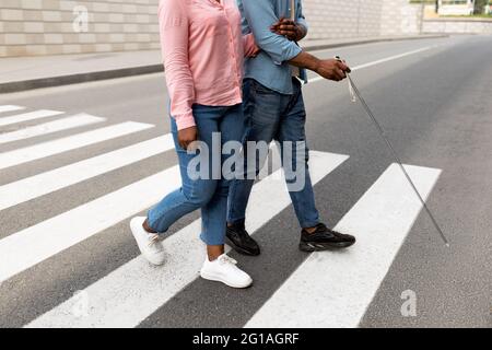 Vue rognée de la femme soignante aidant un homme noir malvoyants avec canne à travers la rue de ville, de plus près Banque D'Images