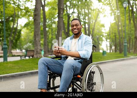 Homme noir handicapé heureux en fauteuil roulant utilisant un smartphone, vérifiant les messages en ligne au parc Banque D'Images