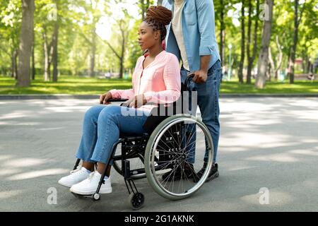 Femme noire handicapée en fauteuil roulant à pied avec son mari affectueux à l'extérieur, passant de grands moments ensemble Banque D'Images