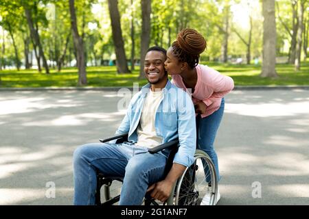 Femme noire affectueuse embrassant son mari handicapé en fauteuil roulant, allant marcher avec lui au parc de la ville Banque D'Images