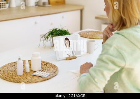 Vue arrière d'une femme d'âge moyen moderne et concentrée ayant un appel en ligne avec un médecin sur un ordinateur portable pendant l'épidémie de Covid 19, assis dans la cuisine à la maison. Concept de santé et de médecine Banque D'Images