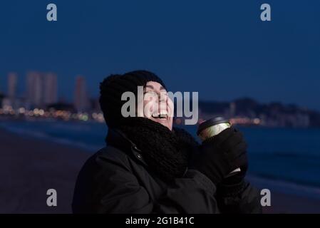 Femme riant à voix haute en marchant sur une nuit froide sur une plage ayant une boisson chaude, vêtue de vêtements sombres et de chapeau, la ville et ses lumières peuvent être s Banque D'Images