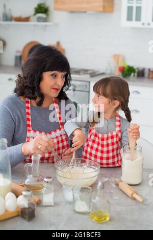 La grand-mère et la petite-fille en tabliers à carreaux cuisent dans la cuisine et se regardent l'une l'autre. Banque D'Images