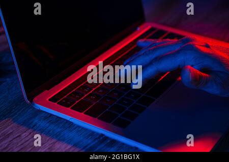 Crop anonyme mâle à l'aide d'un ordinateur portable avec écran noir placé sur une table en bois dans un lieu de travail sombre avec des lumières au néon Banque D'Images