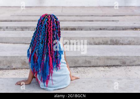 Vue arrière d'un enfant ethnique rêveur méconnaissable avec des tresses colorées, assis sur un escalier en béton tout en regardant loin en plein jour Banque D'Images