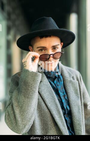 Jeune homme transgenre en manteau classe et chapeau regardant la caméra à la lumière du jour Banque D'Images