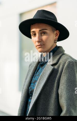 Jeune homme transgenre en manteau classe et chapeau regardant la caméra à la lumière du jour Banque D'Images