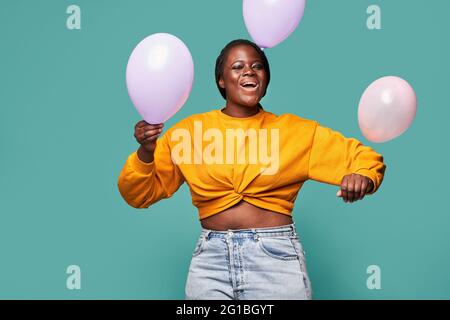 Femme afro-américaine excitée en Jean et jaune debout près de ballons tombant sur fond bleu dans le studio Banque D'Images