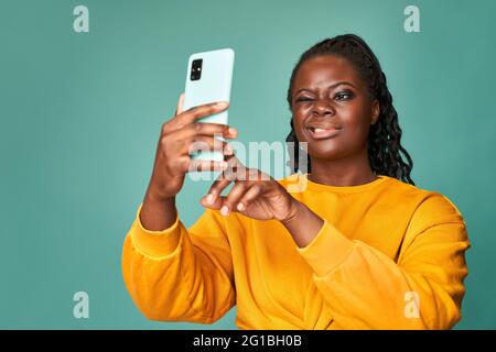 Bonne femme afro-américaine en chandail jaune en train de se tordre et de prendre selfie sur smartphone contre le mur bleu en studio Banque D'Images