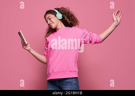 Adolescent plein de gaieté avec ses cheveux bouclés, son bras d'étirement et son téléphone mobile à défilement, tout en écoutant de la musique et en sautant sur fond rose Banque D'Images