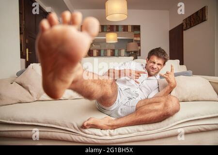 Corps complet de jeune positif barefooted mâle dans des vêtements décontractés atteignant les pieds vers l'appareil photo et signe gestant de cornes tout en se relaxant sur la patte de confort Banque D'Images