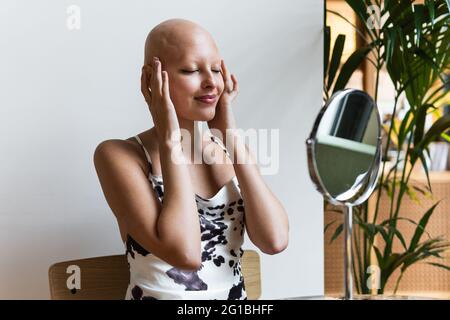 Bonne jeune femme sans cheveux avec la maladie d'alopécie dans une tenue élégante et des lèvres rouges souriant et les yeux fermés devant un petit miroir rond assis sur la cha Banque D'Images