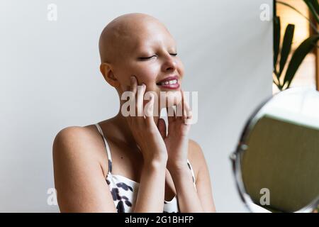 Bonne jeune femme sans cheveux avec la maladie d'alopécie dans une tenue élégante et des lèvres rouges souriant et les yeux fermés devant un petit miroir rond assis sur la cha Banque D'Images