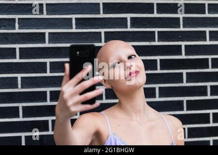 Ravie jeune femme sans cheveux en haut clair avec alopécie maladie auto-immune debout près du mur de brique noire et souriant tout en prenant autoportrait sur les s. Banque D'Images
