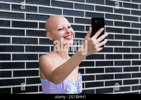 Ravie jeune femme sans cheveux en haut clair avec alopécie maladie auto-immune debout près du mur de brique noire et souriant tout en prenant autoportrait sur les s. Banque D'Images