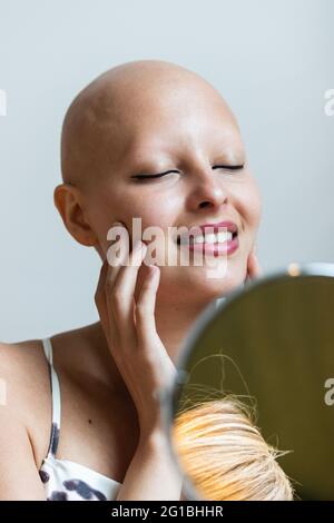 Bonne jeune femme sans cheveux avec la maladie d'alopécie dans une tenue élégante et des lèvres rouges souriant et les yeux fermés devant un petit miroir rond assis sur la cha Banque D'Images