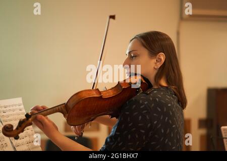 Vue latérale d'une musicienne professionnelle concentrée jouant du violon acoustique et regardant une feuille de musique pendant la répétition en studio Banque D'Images