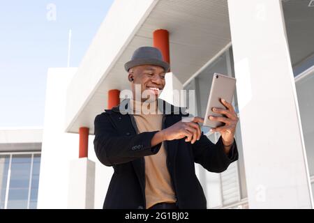 Joyeux Afro-américain dans des vêtements élégants et des écouteurs naviguant sur une tablette moderne dans une rue ensoleillée et regardant l'écran avec le sourire Banque D'Images