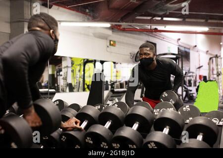 Un jeune sportif afro-américain plein de confiance en matière d'activités et de masque levant de lourdes haltères tout en étant assis sur le banc dans une salle de gym moderne Banque D'Images