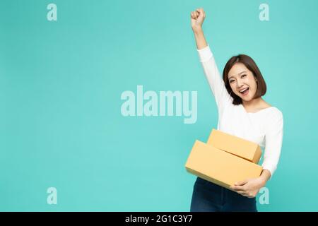 Bonne femme asiatique tenant la boîte colis et célébrant le succès avec les mains dans l'air isolé sur fond vert clair, livraison de courrier et SH Banque D'Images