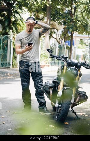 Homme confus avec une clé dans les mains essuyant la sueur de son front et lisant l'article sur la réparation de moto sur son smartphone Banque D'Images