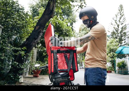 Messagerie dans un casque et un chauffe-cou prenant la commande de nourriture emballée hors du sac thermique pour l'apporter au client Banque D'Images