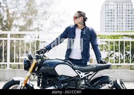 Homme sérieux dans des lunettes de soleil debout à moto et regardant loin Banque D'Images