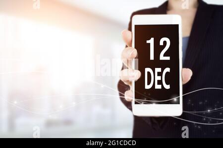 12 décembre. Jour 12 du mois, date du calendrier. Smartphone blanc avec date de calendrier en femme d'affaires main sur fond flou. Mois d'hiver, jour de t Banque D'Images