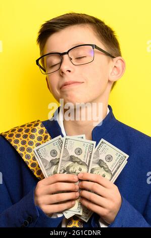 Un garçon en lunettes et un costume surdimensionné avec un visage satisfait tient des dollars US, un enfant et de l'argent gros. Nouveau Banque D'Images