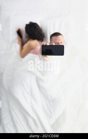 Une femme qui se détourne masque l'écran de son smartphone à son mari pendant qu'il est allongé sur son lit, son mari la regarde à l'aide de l'application de son smartphone Banque D'Images