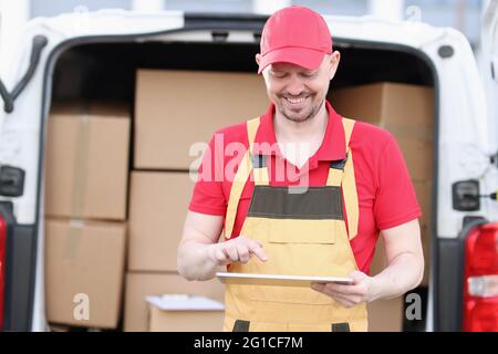 Le joli postier caucasien en costume et casquette vérifie les boîtes aux lettres avec un appareil de tablette dans ses mains Banque D'Images