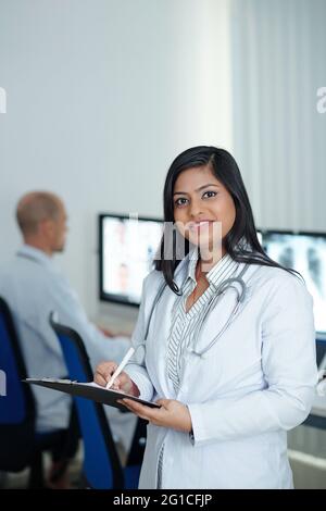 Portrait d'une jeune femme médecin positive prenant des notes dans le document après avoir eu une réunion en ligne avec des collègues d'autres cliniques Banque D'Images