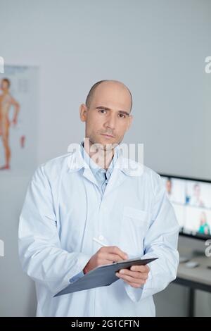 Portrait d'un médecin généraliste sérieux en blouse de travail blanche écrivant les données des patients dans le document Banque D'Images