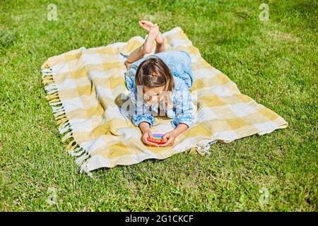 Une fille se trouve sur une couverture d'herbe à l'extérieur et joue pop il, les mains d'enfants jouant des bulles d'un jouet étriquée arc-en-ciel, jouet de violon dans l'arrière-cour du ho Banque D'Images