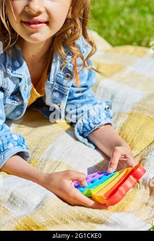 Une fille se trouve sur une couverture d'herbe à l'extérieur et joue pop il, les mains d'enfants jouant des bulles d'un jouet étriquée arc-en-ciel, jouet de violon dans l'arrière-cour du ho Banque D'Images