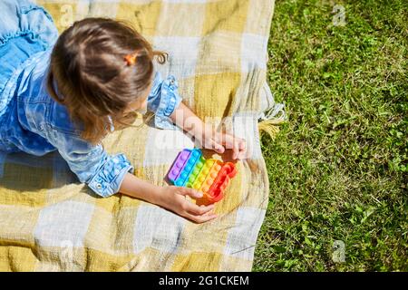 Une fille se trouve sur une couverture d'herbe à l'extérieur et joue pop il, les mains d'enfants jouant des bulles d'un jouet étriquée arc-en-ciel, jouet de violon dans l'arrière-cour du ho Banque D'Images