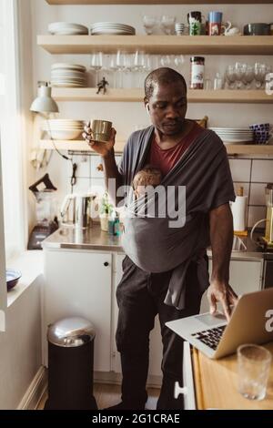 Homme d'affaires avec son fils dans un porte-bébé utilisant un ordinateur portable tout en tenant une tasse dans la cuisine Banque D'Images