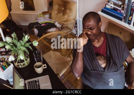 Un père fatigué avec un bébé dans un porteur assis au bureau à domicile Banque D'Images