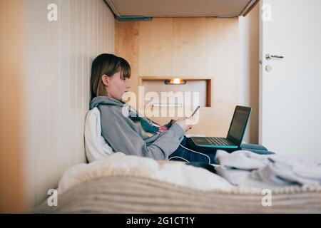 Une adolescente assise avec un ordinateur portable à l'aide d'un smartphone dans une maison de vacances Banque D'Images