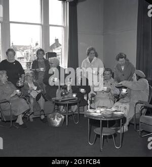 Années 1960, historique, à l'intérieur d'une salle de séjour dans une maison de soins, un groupe de femmes âgées ayant du thé et des biscuits, assis dans des chaises autour d'une théière plaquée en métal, sur un stand en bois, enfermé « The Singing Kettle Club », Angleterre, Royaume-Uni. Banque D'Images